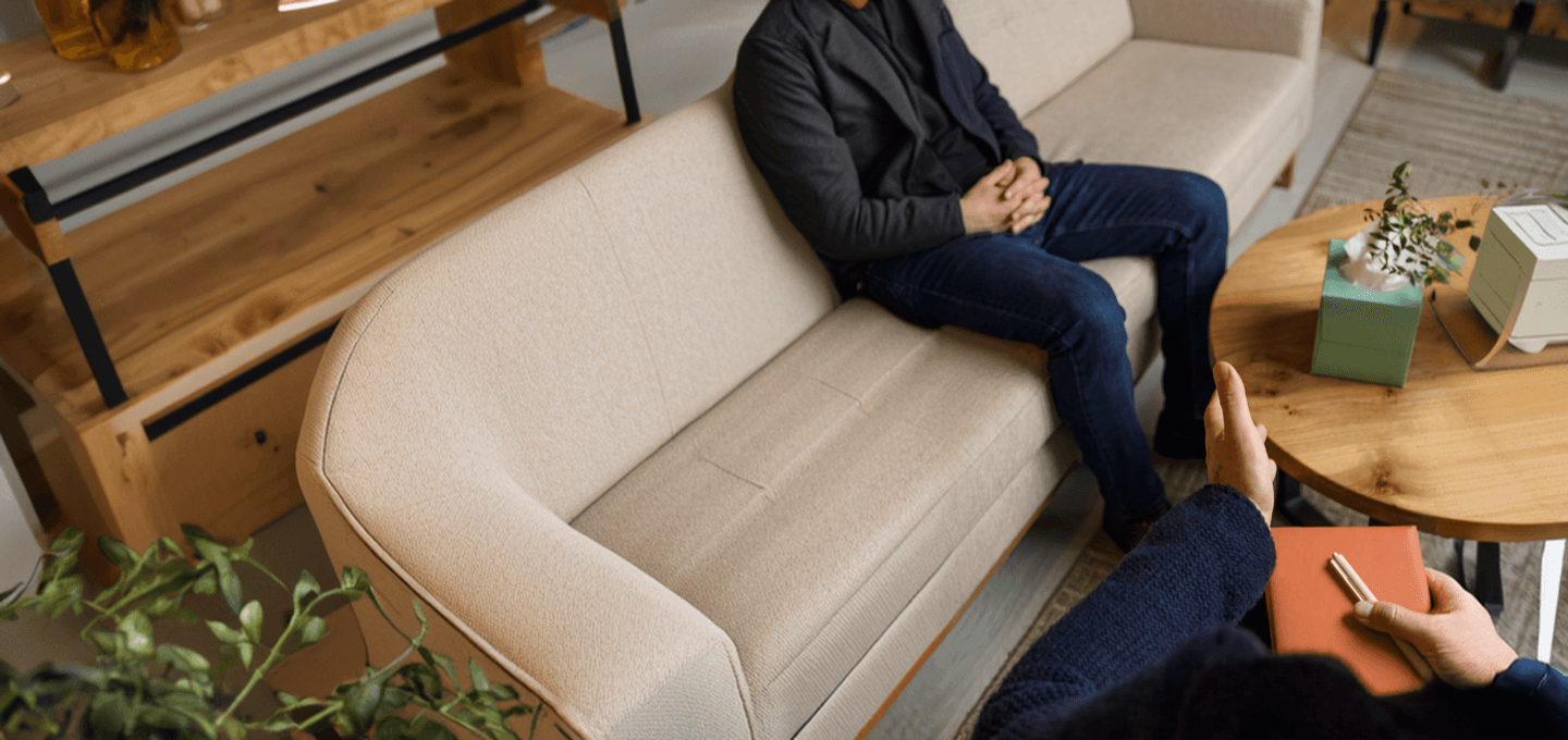 A man sitting on a couch in a warm therapy office while a clinician takes notes