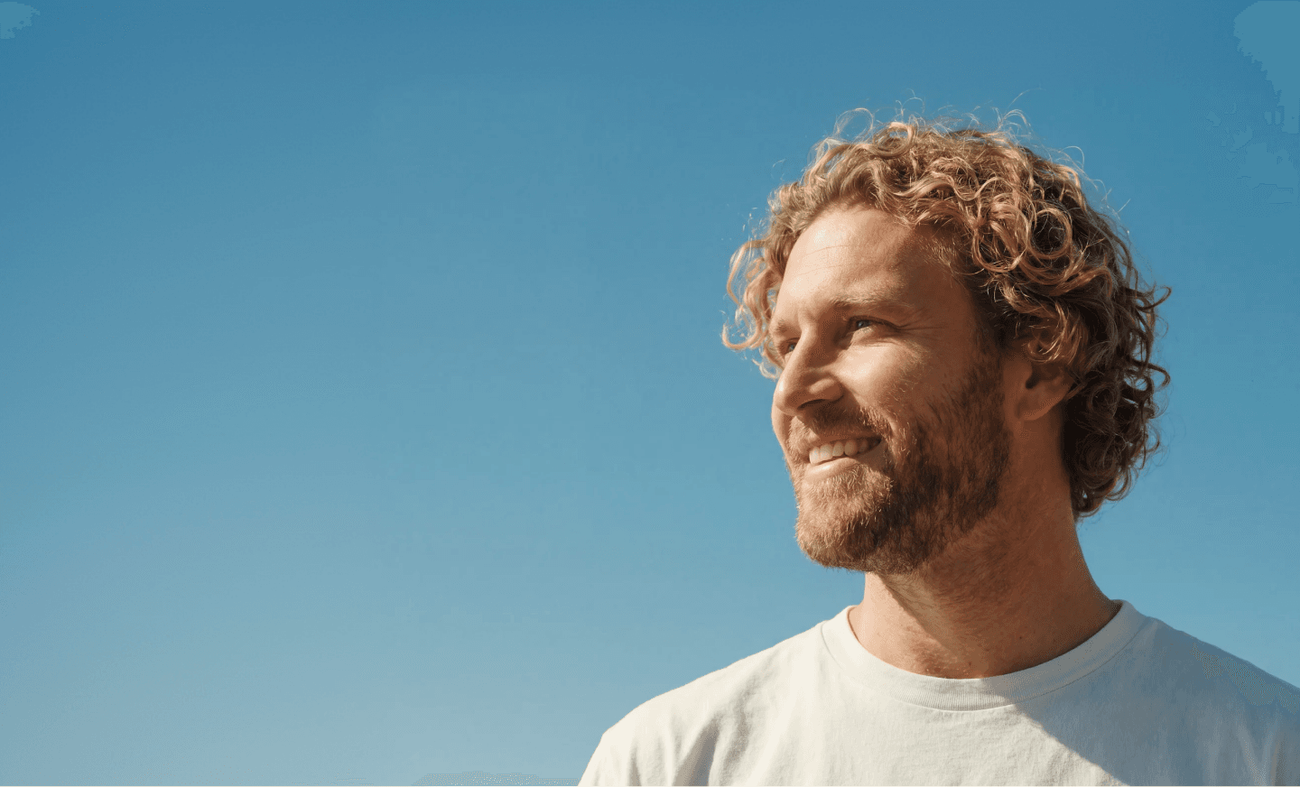 A smiling man with curly hair looking up against a clear blue sky