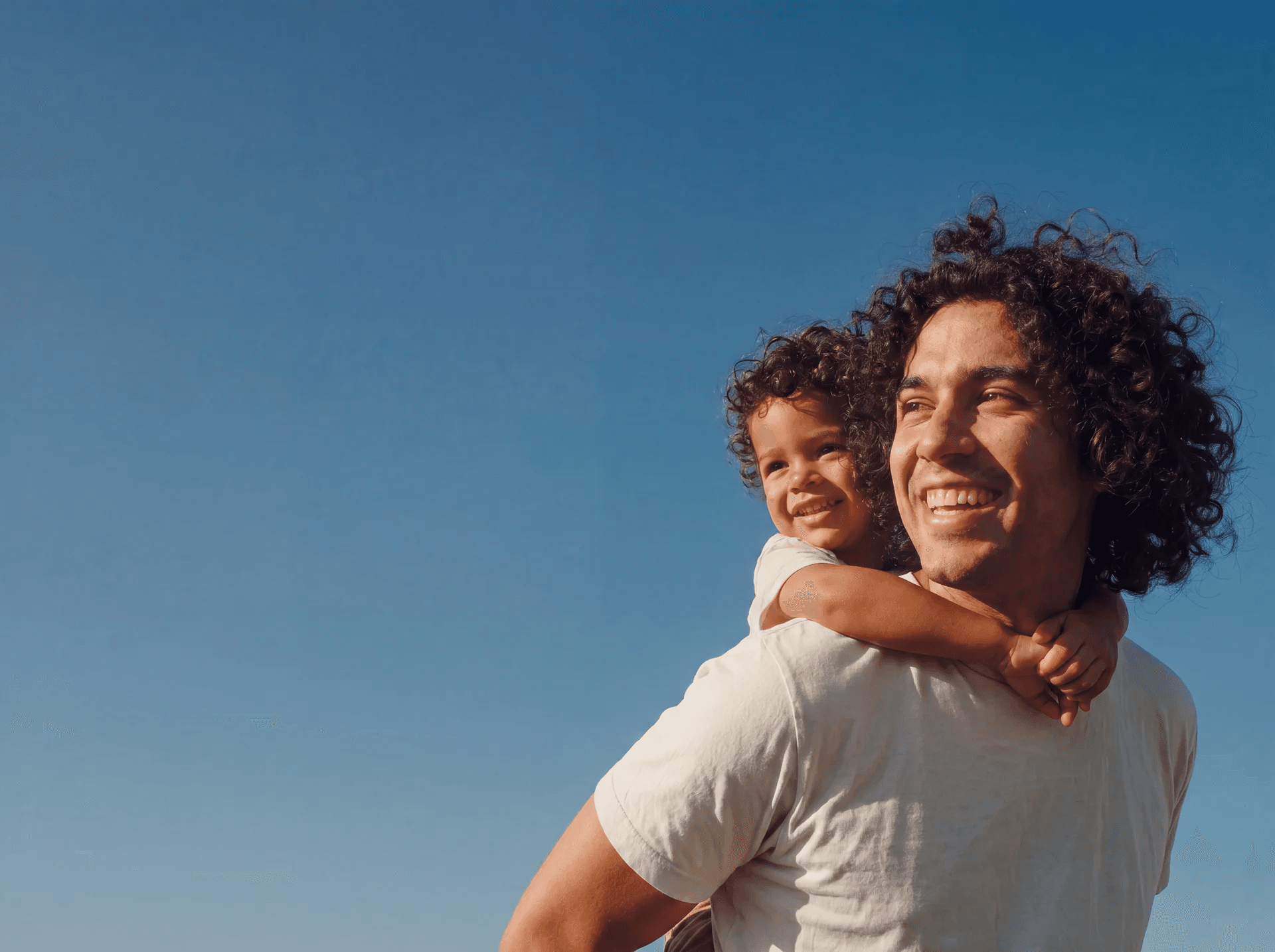 A father carrying his laughing toddler on his shoulders against a clear blue sky
